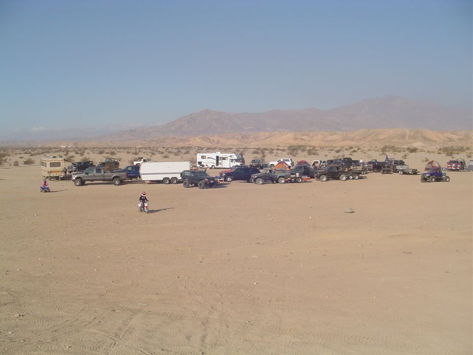 Desert scene with numerous off-road vehicles, trucks, and RVs parked on sandy terrain under a clear blue sky