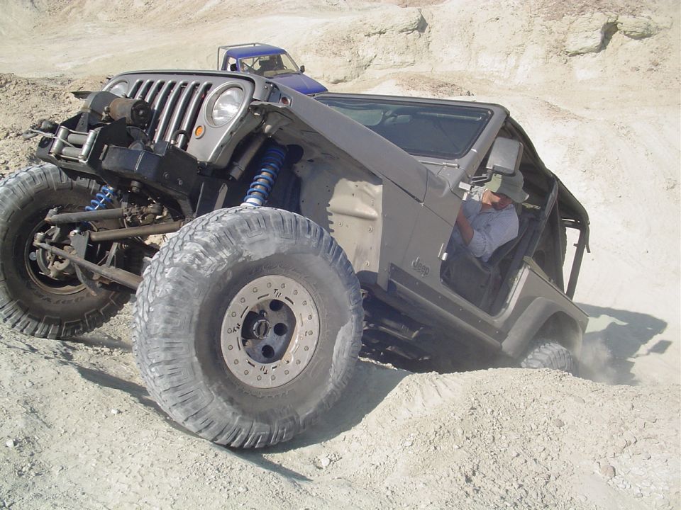 Modified Jeep TJ with large tires and suspension, navigating rough terrain. Another blue vehicle is visible in the background