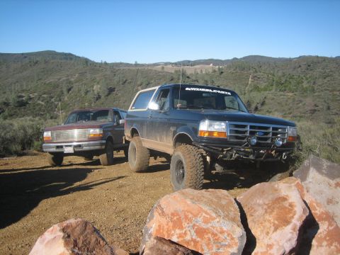 1995 Ford Bronco - Two Ford Broncos are seen off-roading on a sunny day with hills in the background.