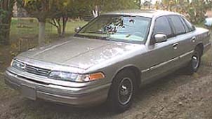 1994 Ford Crown Victoria - A silver 1994 Ford Crown Victoria is parked on a dirt lot with trees in the background.