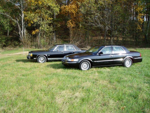Two black sedans parked on grass, one in front of the other. The front car is a 1998 Mercury Grand Marquis
