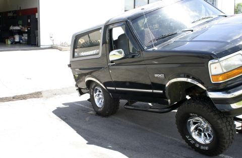 1994 Ford Bronco - A black Ford Bronco is parked outside a garage. The vehicle has chrome accents and off-road tires.