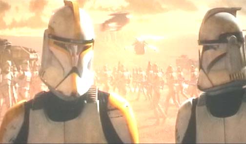Two clone troopers in helmets stand in a desert landscape with a crowd and vehicles in the background