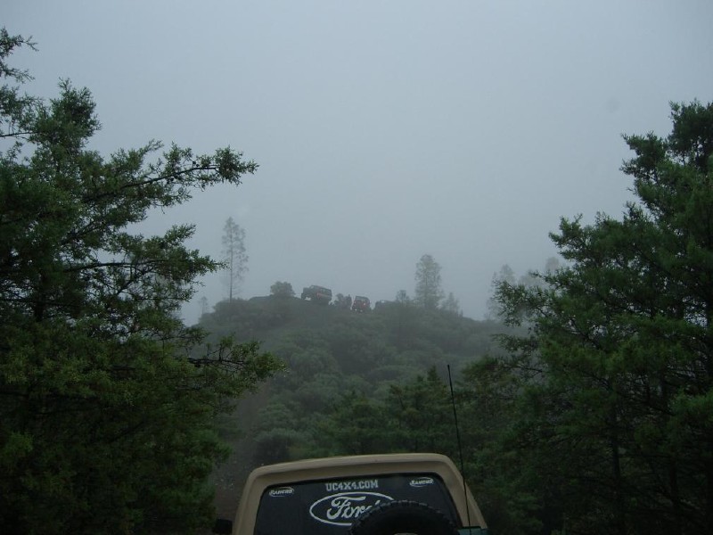 Several off-road vehicles drive up a foggy hill, seen from the back of another vehicle