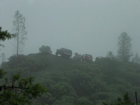 1989 Ford Bronco - Multiple SUVs navigate a misty, wooded incline, with the rear of the lead vehicle illuminated by its taillights.