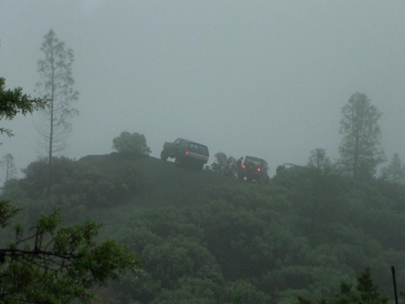 Three dark SUVs drive up a foggy, tree-covered hill. The lead vehicle's taillights are visible