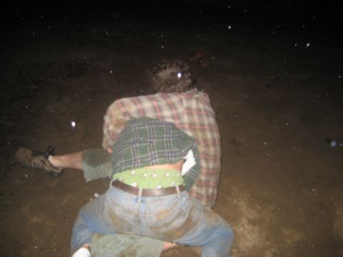 1966 Jeep CJ5 - Two individuals are seen from behind in a muddy outdoor environment at night, with one person wearing a green plaid shirt over a tan and re…