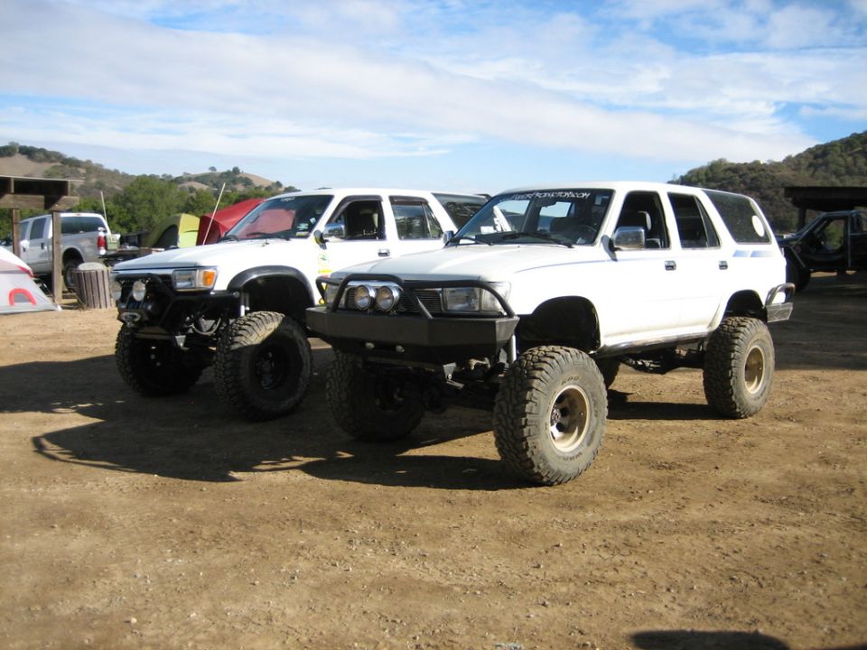 Two white Toyota 4Runners parked on dirt, both lifted with large tires. One has a custom bumper
