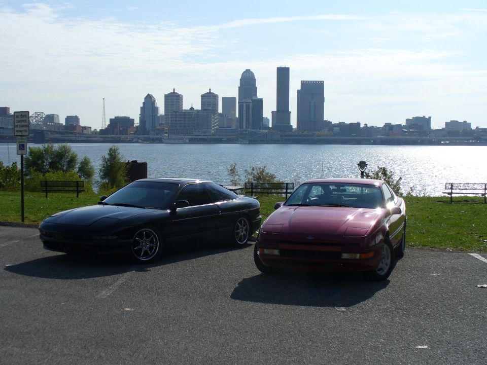 Two 1991 Ford Probes parked by a river with a city skyline