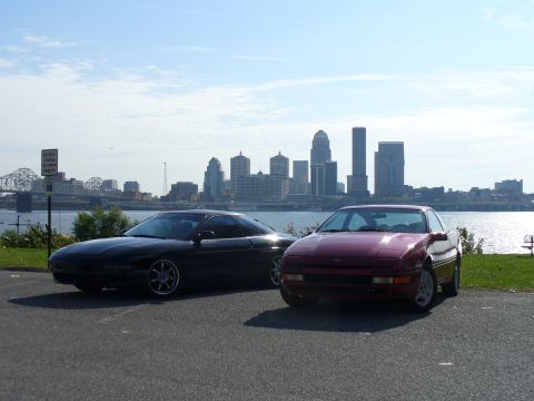 1991 Ford Probe - Two Ford Probe hatchback coupes parked side by side on a riverside lot, a black second-generation car on chrome multi-spoke wheels and a ma…