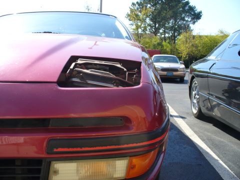 1991 Ford Probe - Tight, low-angle shot down the nose of a maroon Ford Probe with the passenger-side pop-up headlamp bucket removed, exposing the empty housi…