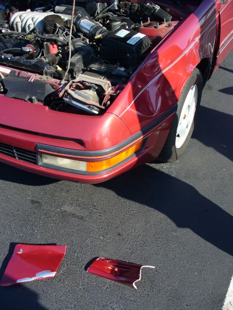 1991 Ford Probe - Front view of a 1991 Ford Probe with the hood up, revealing engine components and broken bumper parts on the asphalt.