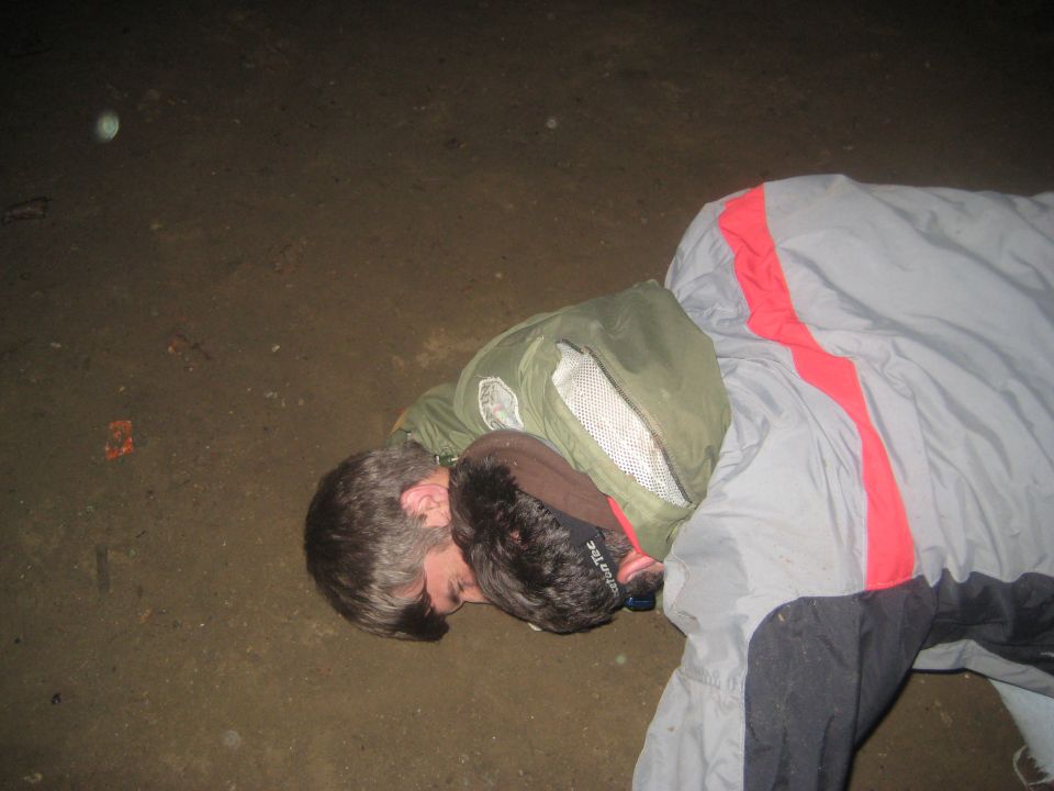 Two people sleeping on the ground, one in a green jacket and gray sleeping bag with red stripe