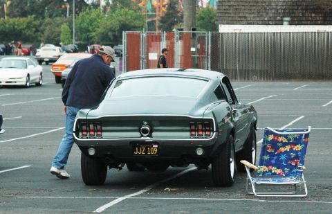 1991 Ford Taurus - A man inspects a classic Ford Mustang Fastback parked in a lot, with a beach chair set up next to it.