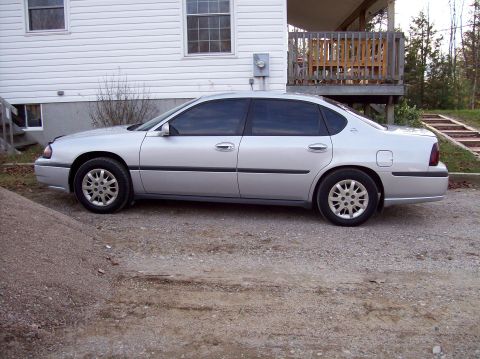 2001 Chevrolet Impala - Side view of a silver sedan parked on a gravel driveway next to a house.