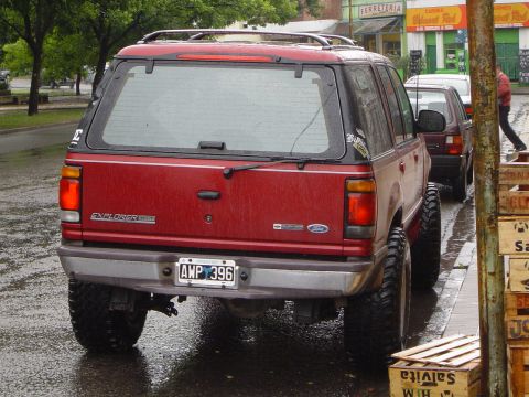 1996 Ford Explorer - A red 1996 Ford Explorer is parked on a wet street, a lifted suspension and large off-road tires. Another vehicle is visible behind it.