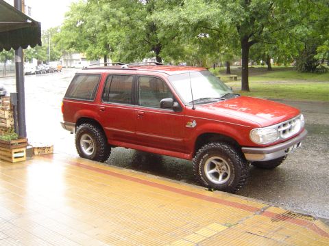1996 Ford Explorer - Red Ford Explorer SUV with lifted suspension and off-road tires parked on a wet sidewalk.