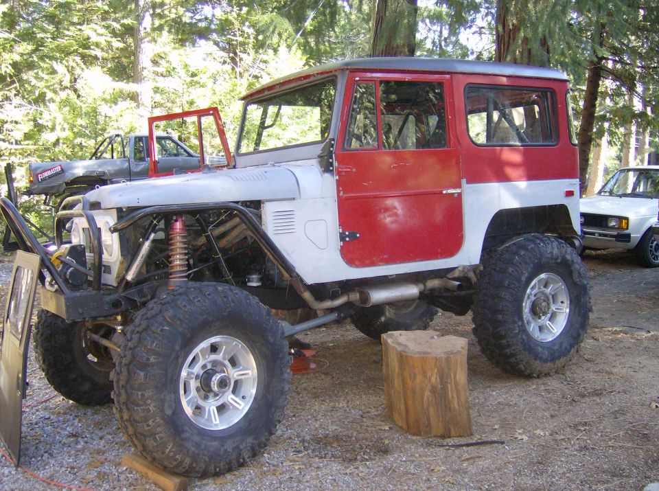 Modified Toyota Land Cruiser FJ40 with red doors and silver body, lifted with large tires. Another truck and a white car are in the background