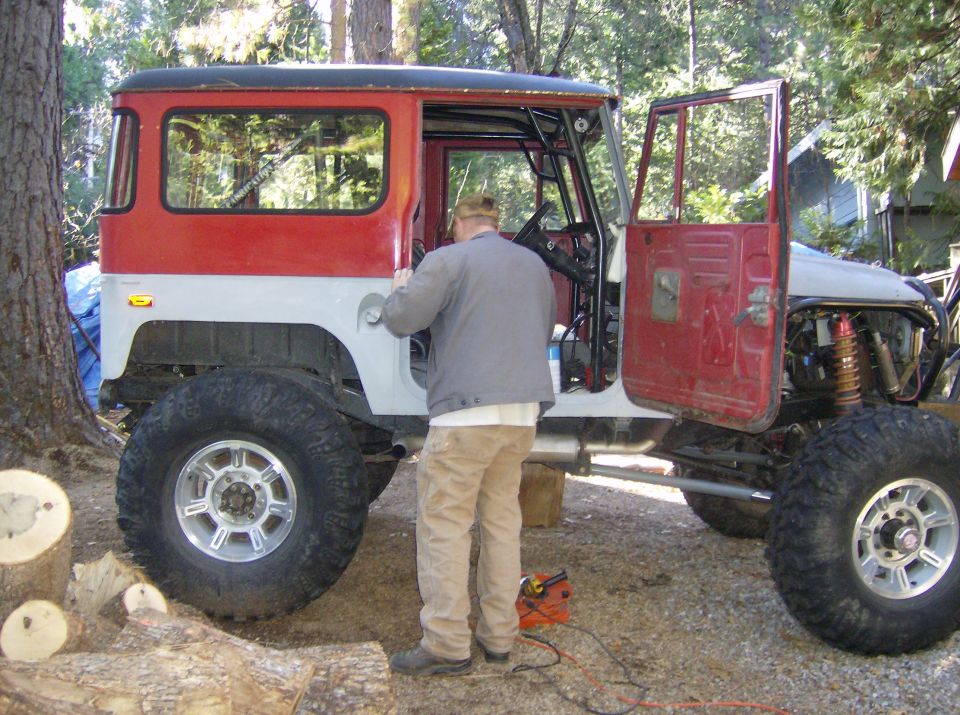 Red and white Toyota Land Cruiser FJ40 with large tires and roll cage, driver's door open, man working on it