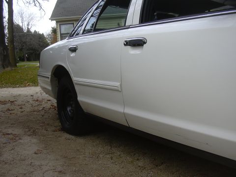 1997 Ford Crown Victoria - The side profile of a white 1997 Ford Crown Victoria is visible, parked outdoors on a dirt surface.