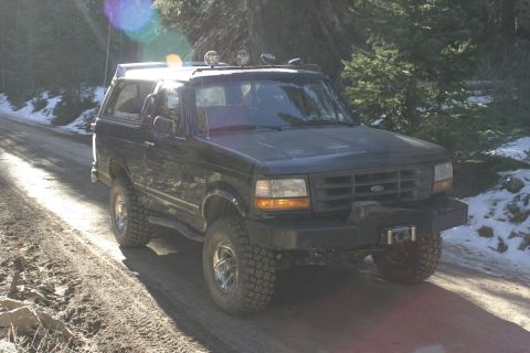 1994 Ford Bronco - A black Ford Bronco is parked on a dirt road with trees and snow in the background.