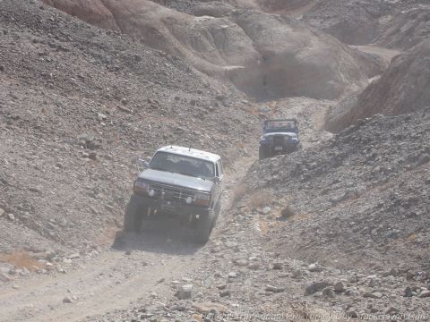 1994 Ford Bronco - Two off-road vehicles navigate a steep, rocky path in a desert canyon.