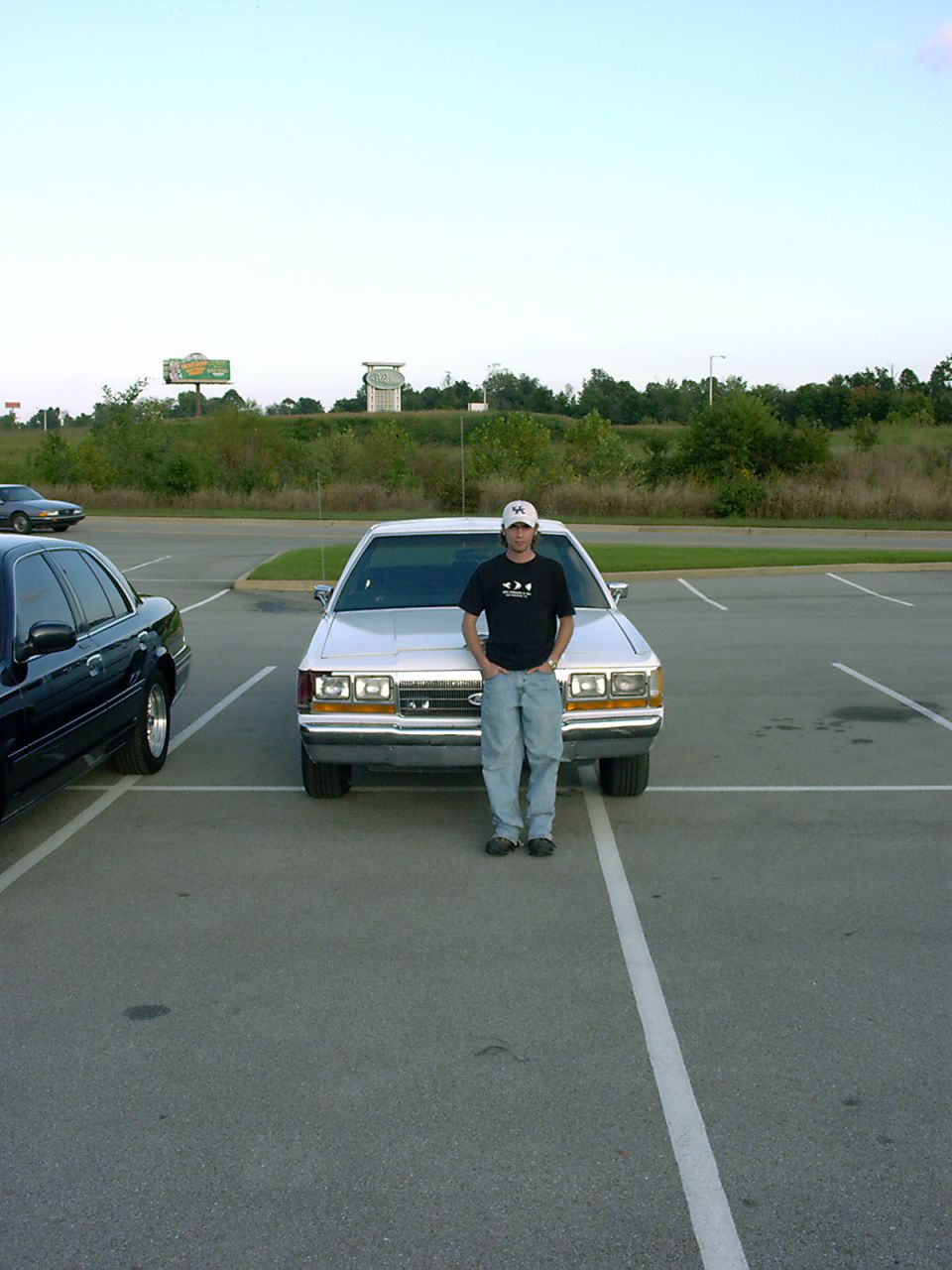 Man in cap standing in front of silver Ford LTD Crown Victoria with woodgrain trim