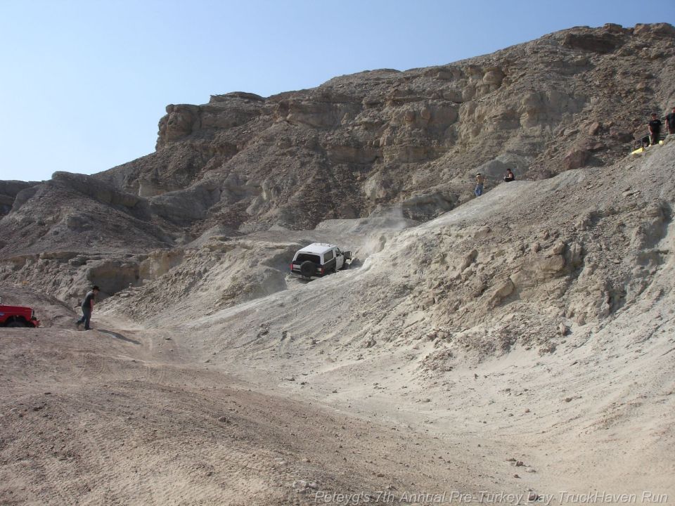 White SUV driving down a steep, dusty, rocky hill with a red vehicle parked nearby