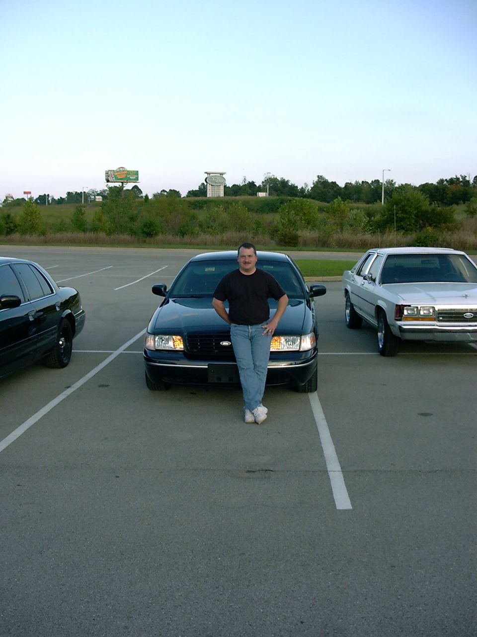 Man standing in front of dark Ford Crown Victoria sedan in parking lot at dusk