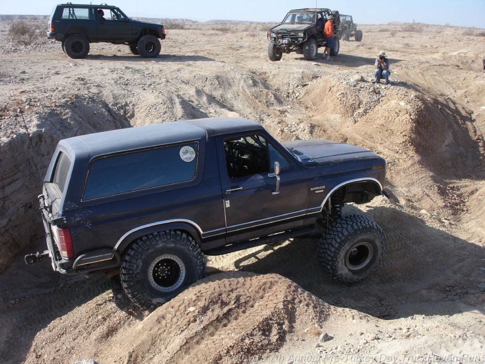 Dark blue Ford Bronco with lifted suspension navigating a dirt obstacle, other off-road vehicles and people in background