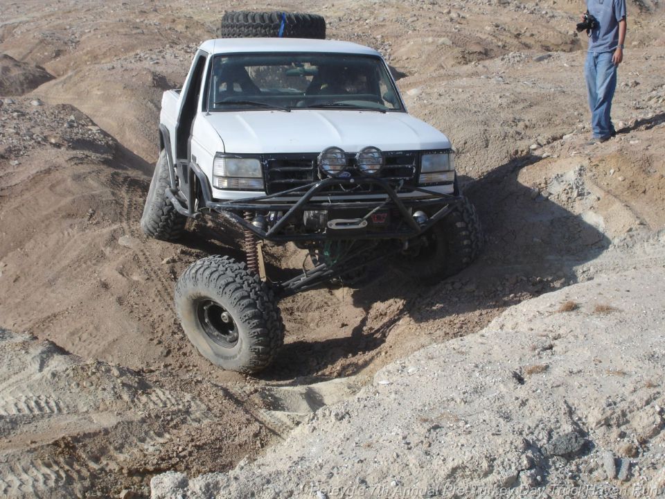 White pickup truck with off-road modifications climbing a dirt hill. Spare tire on roof