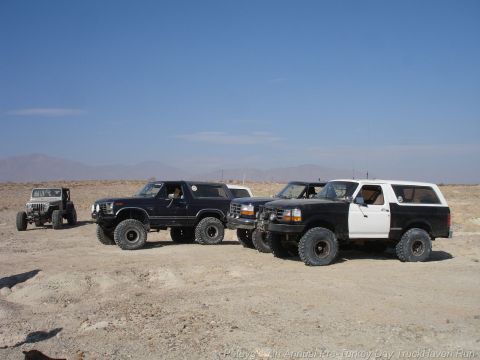 1994 Ford Bronco - Multiple Broncos and a Jeep Wrangler are parked in a desert area under a clear blue sky.