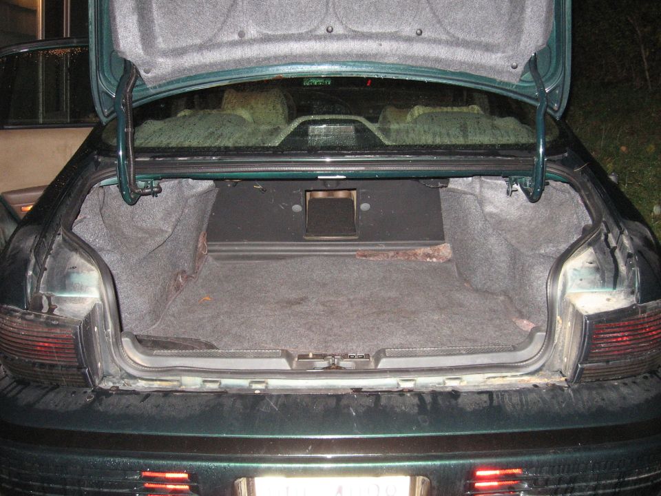 Open trunk of a dark green car showing carpeted interior and a black speaker box mounted in the center