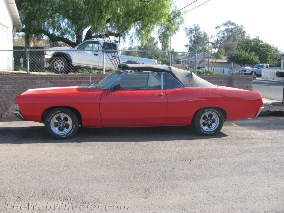 Side profile of a red convertible with black soft top raised, parked on gravel