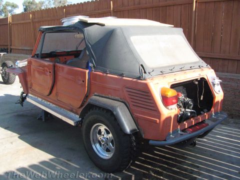 1990 Ford Bronco - orange Volkswagen Thing with its soft top up and rear engine compartment open, parked on pavement against a wood fence.