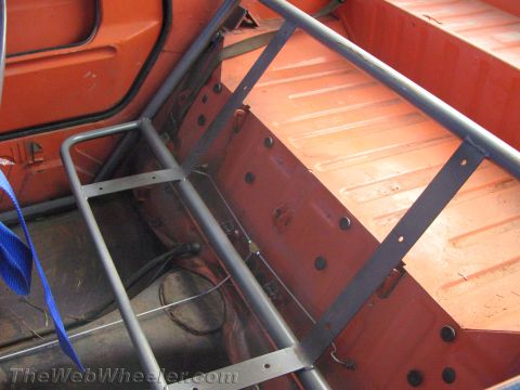 1990 Ford Bronco - Bare orange floor pan and frame of a vehicle with a gray tubular roll cage being fabricated and a blue tie-down strap visible.