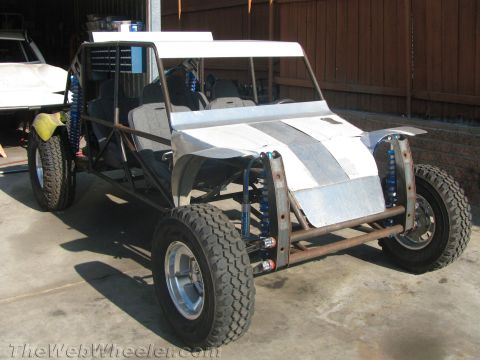 1990 Ford Bronco - tube-frame off-road sand rail buggy with blue coilover shocks and bare aluminum bodywork parked in a garage.