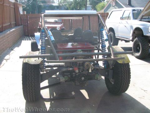1990 Ford Bronco - Rear view of a tube-frame off-road buggy revealing its rear axle, driveshaft, and blue coilover shocks, with a white SUV nearby.