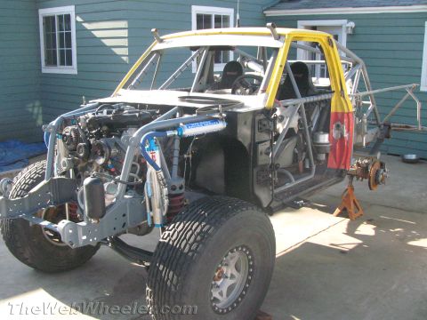 1990 Ford Bronco - stripped tube-chassis race truck on jack stands showing its engine, long-travel suspension, and bare frame beside a green house.