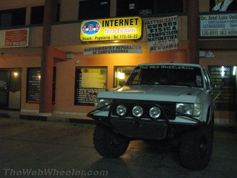 1990 Ford Bronco - white off-road truck with a front light bar parked at night outside an illuminated S.O.S Internet storefront and signage.