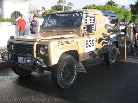 1990 Ford Bronco - tan Land Rover Defender race truck numbered 839 with flame graphics and BFGoodrich tires parked on a street with onlookers.