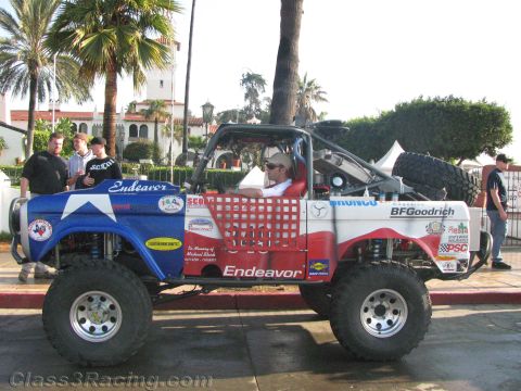 1990 Ford Bronco - Profile of a heavily lifted early Ford Bronco off-road racer in patriotic Endeavor Racing livery, driver seated, parked on a palm-lined str…