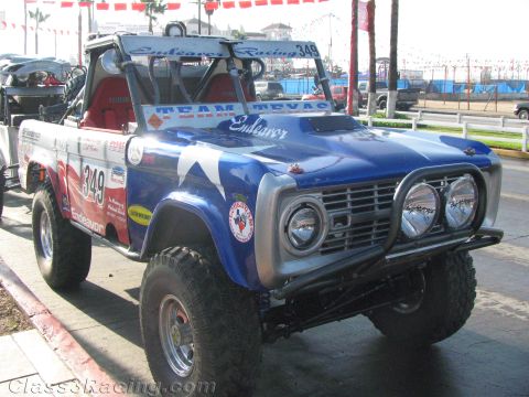 1990 Ford Bronco - Front three-quarter view of a lifted early Ford Bronco off-road racer #349 with triple headlights and a tubular brush guard at a staging ar…