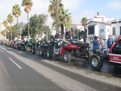 1990 Ford Bronco - line of single-seat off-road race buggies and ATVs staged curbside along a palm-lined wet street before an event.