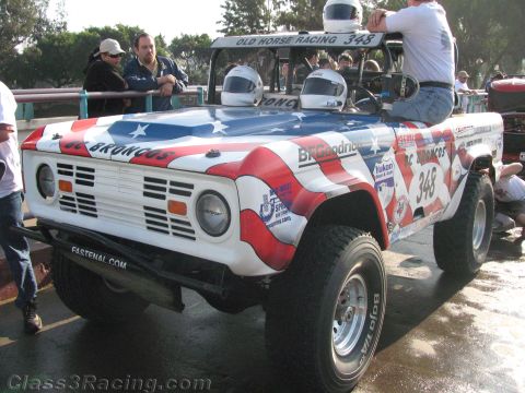 1990 Ford Bronco - Front three-quarter view of an early Bronco off-road racer #348 in an American-flag wrap with helmeted occupants in the open cab.