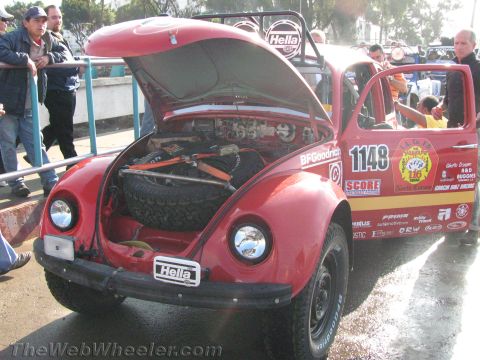 1990 Ford Bronco - red Baja-prepped Volkswagen Beetle racer #1148 with its front trunk open showing a spare tire, parked among a crowd.