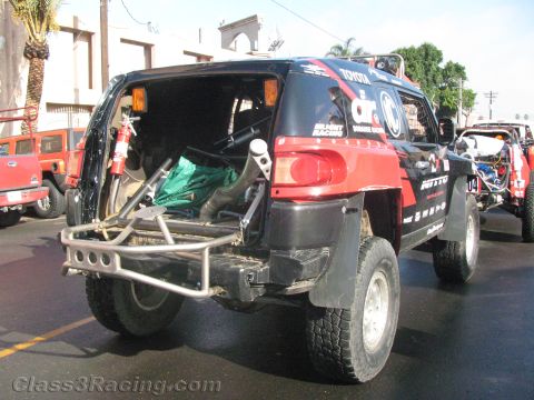 1990 Ford Bronco - Rear three-quarter view of a black and red Toyota FJ Cruiser off-road racer with its rear door open showing cargo and a tubular bumper.