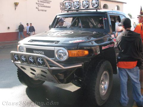 1990 Ford Bronco - Front three-quarter view of a black Toyota FJ Cruiser off-road racer with a roof light bar and front-mounted spotlights at a street staging…