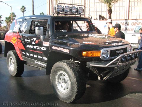 1990 Ford Bronco - Front three-quarter view of a black and red Toyota FJ Cruiser off-road racer in Nitto livery with a roof light bar and tube bumper.