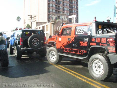 1990 Ford Bronco - black Hummer H3 beside a red and black Hummer H2 off-road race truck #866 staged on a city street near tall buildings.